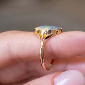 A close-up of an antique, Edwardian era opal solitaire ring with decorative split shoulders mounted in yellow gold seen on a finger to display the band and its details.