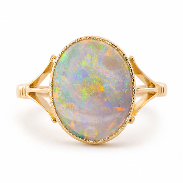 An Antique, Edwardian era oval opal solitaire ring with split yellow gold shoulders and a decorative rub-over setting shown against a white background.