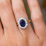 A close-up of a hand wearing a Vintage 1940s cluster engagement ring set to centre with an oval-cut deep blue sapphire surrounded by a halo of diamonds, set on a yellow gold band. 