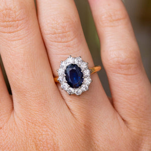 A close-up of a hand wearing a Vintage 1940s cluster engagement ring set to centre with an oval-cut deep blue sapphire surrounded by a halo of diamonds, set on a yellow gold band. 