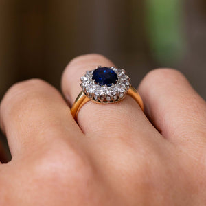A close-up of a Vintage, 1940s yellow gold engagement ring, showcasing the large blue oval central sapphire and surrounding cluster of diamonds.  Worn on a persons finger against a softly blurred background. 