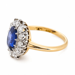 A Vintage, 1940s oval blue sapphire and diamond cluster engagement ring, set on an angle to showcase the side profile and yellow gold band, shown on a plain white background.
