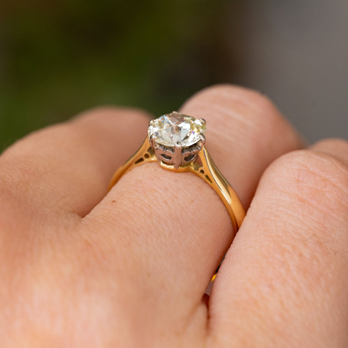 A close-up shot of a yellow gold Antique, Edwardian yellow tinted diamond solitaire engagement ring seen from a low angle to display the gallery worn on a person's finger against a blurred green and grey background.