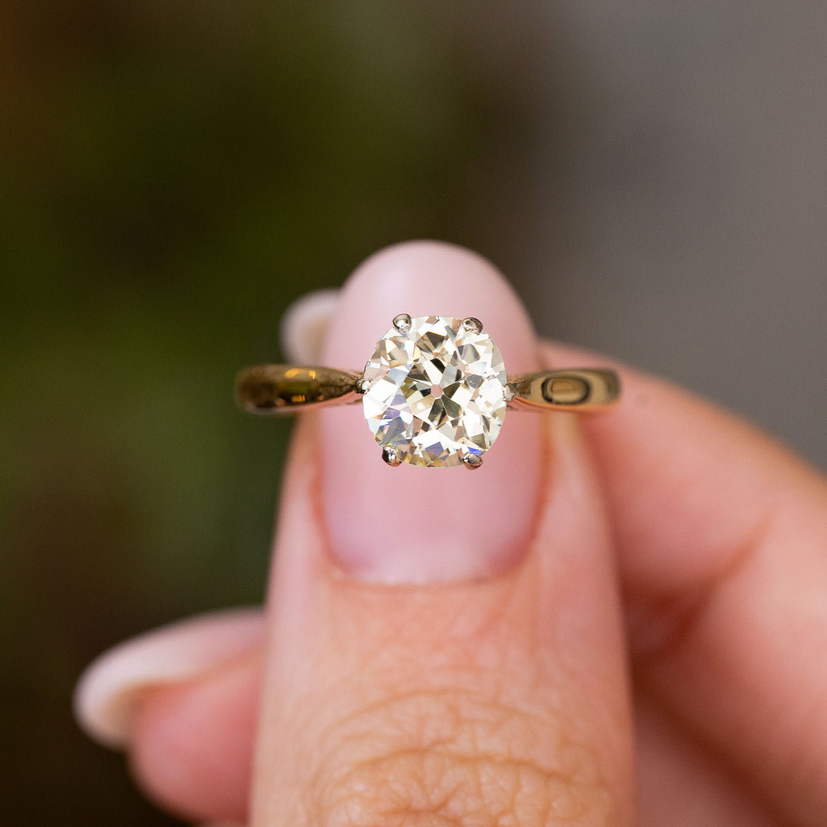 A yellow gold Antique, Edwardian yellow tinted diamond solitaire engagement ring held on a finger against a blurred background
