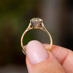 A close-up of a yellow gold Antique, Edwardian yellow tinted diamond solitaire engagement ring held between fingers against a blurred background, held up to show the ring's profile and band.