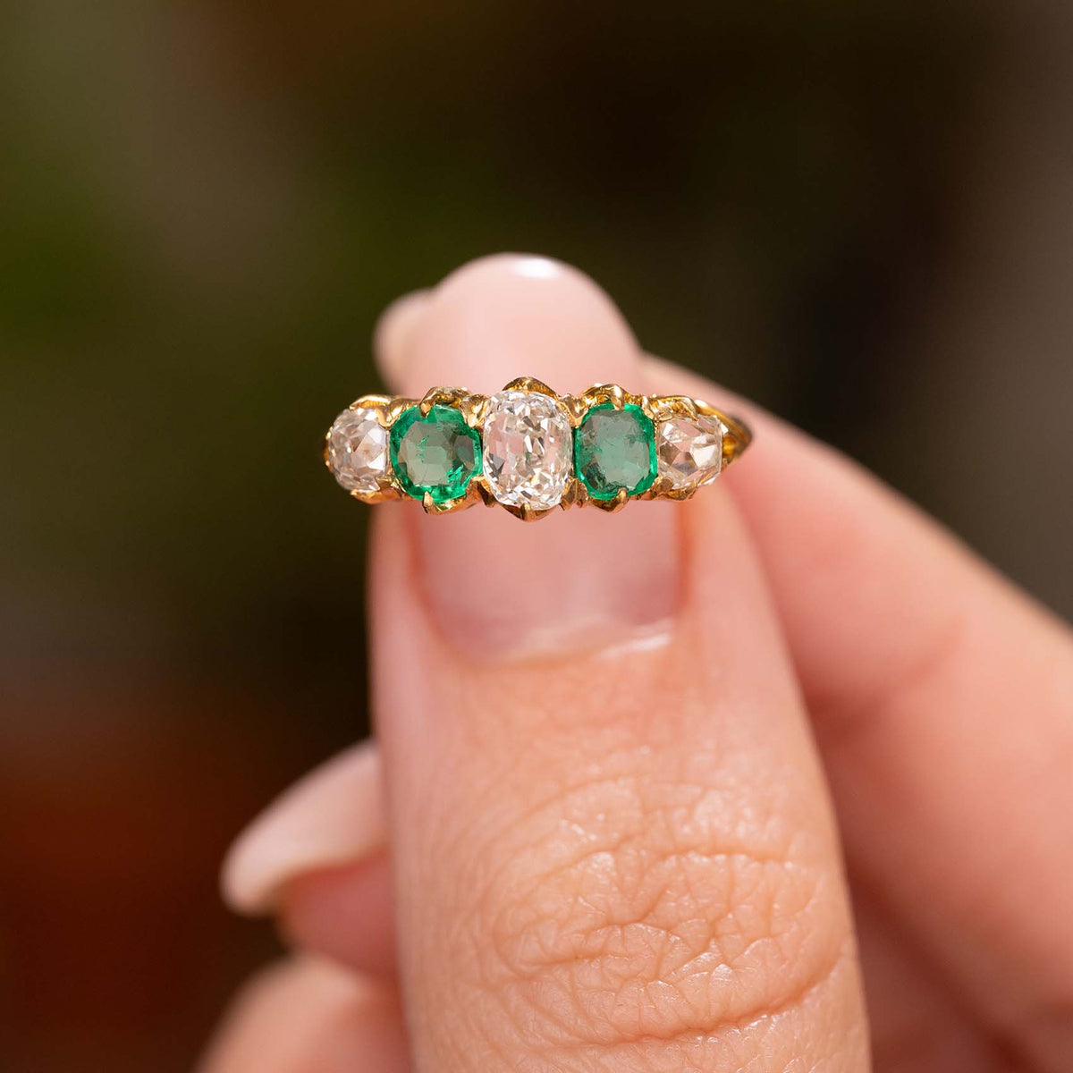 A close-up of a gold ring with two emeralds and three old-cut diamonds, held between fingers against a blurred dark background.