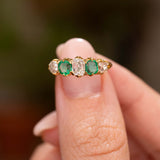 A close-up of a gold ring with two emeralds and three old-cut diamonds, held between fingers against a blurred dark background.