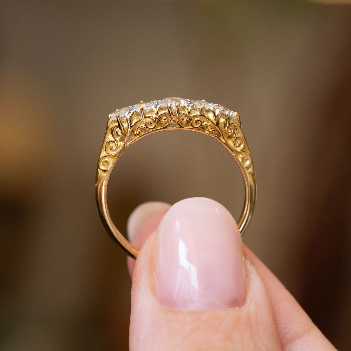 A close-up of an Antique Victorian yellow gold engagement ring with an ornate filigree band and a row of five diamonds, held between fingers against a softly blurred warm brown background.