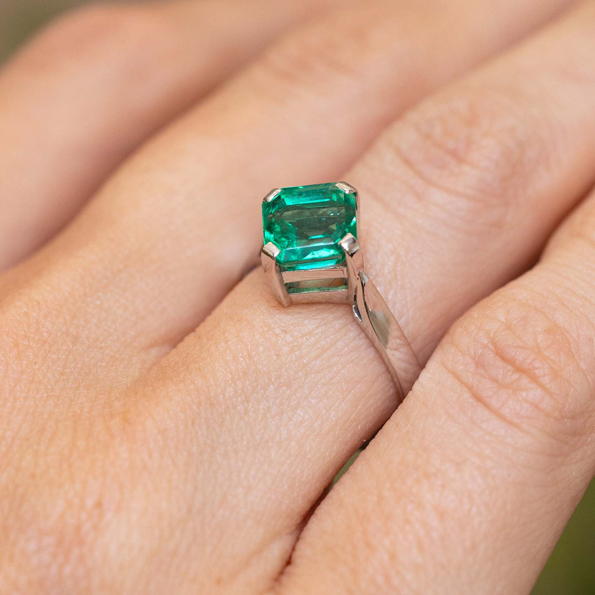 A close up of a square-cut emerald set in a white gold Vintage, 1990s solitaire engagement ring shown worn on a person's finger.