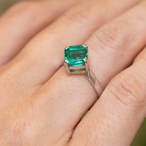 A close up of a square-cut emerald set in a white gold Vintage, 1990s solitaire engagement ring shown worn on a person's finger.