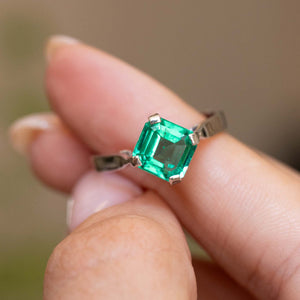 A close-up of a Vintage, 1990s emerald solitaire engagement ring in white gold  worn on a person's finger.