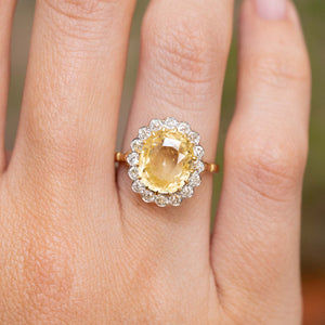 A vintage yellow oval-cut sapphire engagement ring with a gold band and diamonds, shown worn on a person's finger against a softly blurred  background.