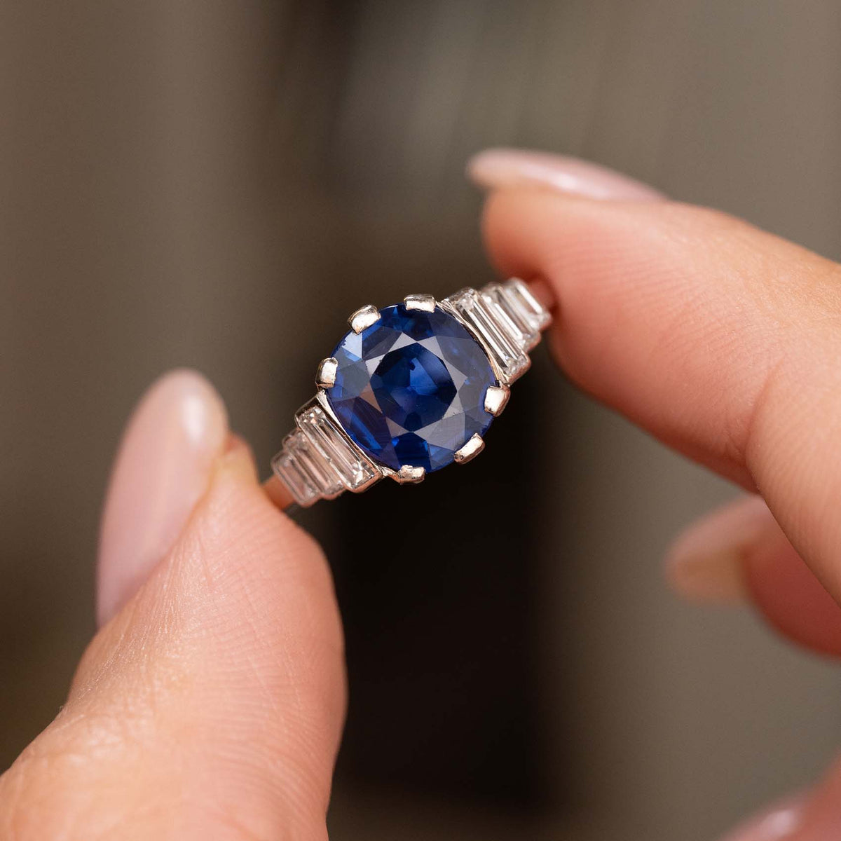 A close-up of a hand holding an Antique, Art Deco blue sapphire and diamond platinum engagement ring against a blurred background.