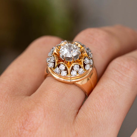 A close-up of a vintage dome cocktail ring from the 1960s set with a large old cut diamond in the centre, surrounded by smaller diamonds and intricate yellow gold work.  Worn on a finger against a blurred background. 