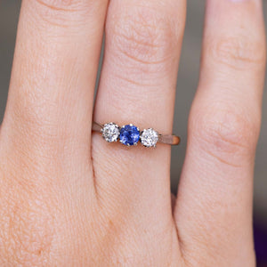 A close-up of an antique trilogy engagement ring worn on a finger, showcasing the central blue sapphire and two diamonds in a platinum setting on a yellow gold band.