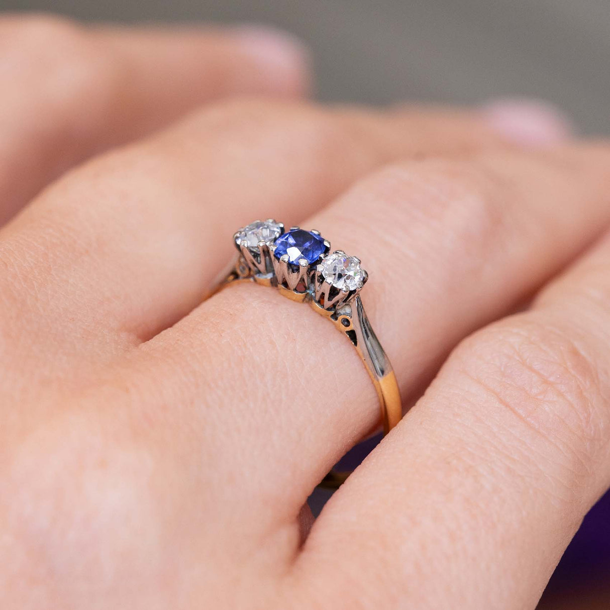 A close-up of an antique trilogy engagement ring in platinum and yellow gold, of a blue sapphire and two diamonds  in a claw setting shown from a side angle to display the gallery.