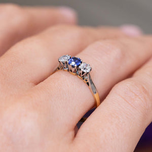 A close-up of an antique trilogy engagement ring in platinum and yellow gold, of a blue sapphire and two diamonds  in a claw setting shown from a side angle to display the gallery.