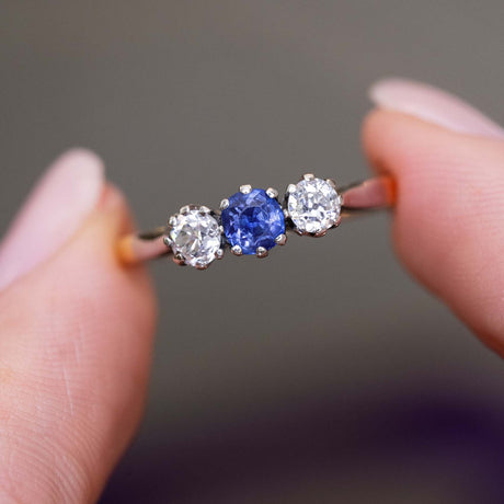 A close-up picture of an antique trilogy engagement ring with platinum settings and a yellow gold band held in between two fingers at a slight angle and shown against a blurred background.