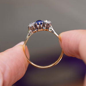 A close up picture of an antique Art Deco trilogy engagement ring with a blue sapphire and two diamonds on a yellow gold band held between the fingers to show the profile of the ring.