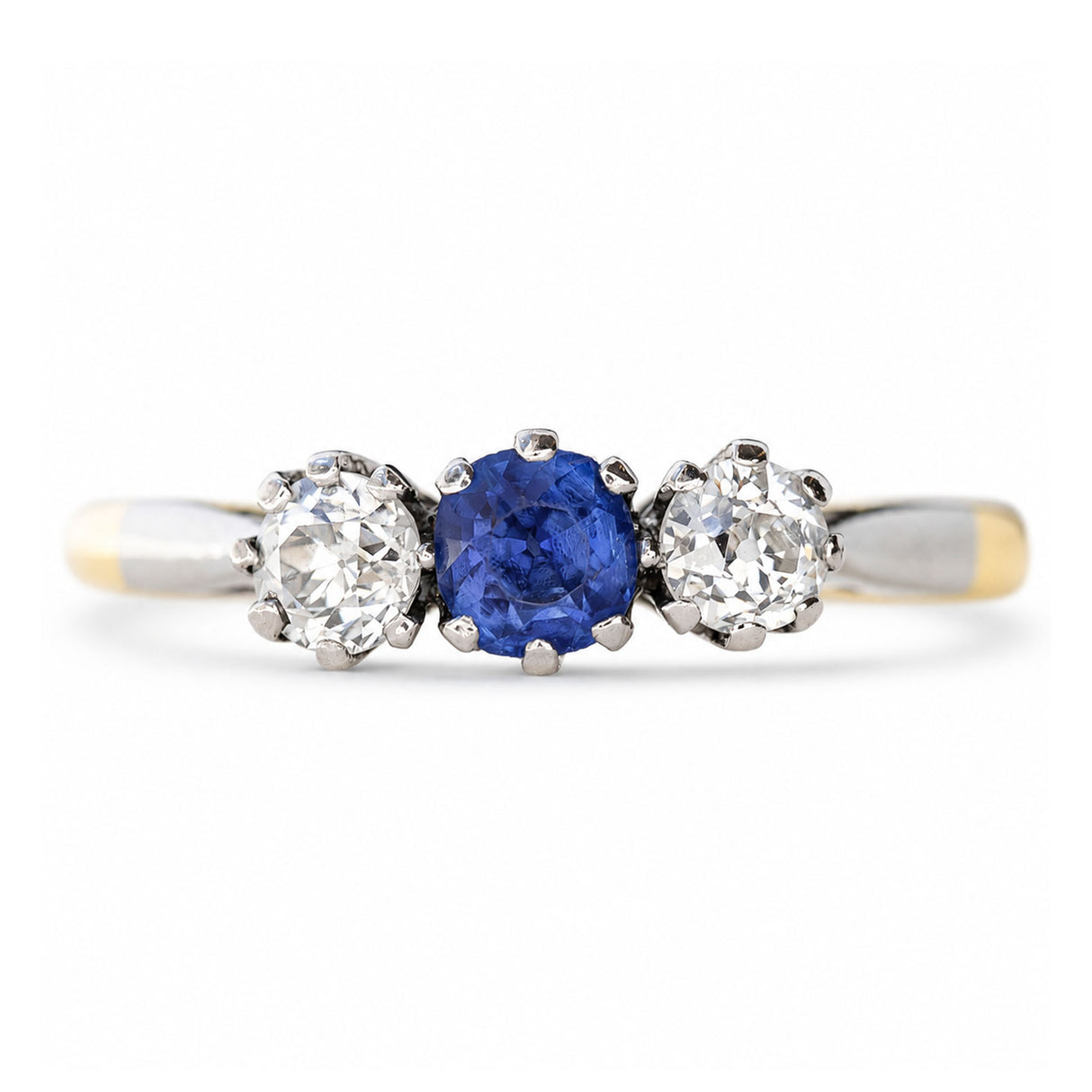 An Antique, Edwardian blue sapphire and diamond three-stone engagement ring in platinum settings on a yellow gold band shown against a white background.