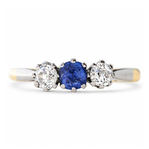 An Antique, Edwardian blue sapphire and diamond three-stone engagement ring in platinum settings on a yellow gold band shown against a white background.