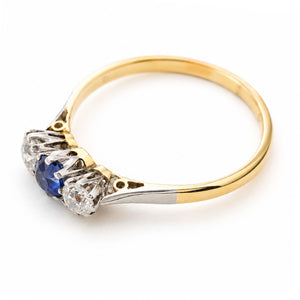 An Antique, Edwardian era blue sapphire and diamond three-stone engagement ring on a yellow gold band shown from above against a white background to showcase the claw-style settings and the details of the shoulders.