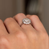 A close-up of an Antique, Edwardian diamond cluster engagement ring in platinum worn on the hand and shown from a low angle to showcase the setting style. 