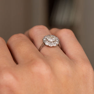 A close-up of an Antique, Edwardian diamond cluster engagement ring in platinum worn on the hand and shown from a low angle to showcase the setting style. 