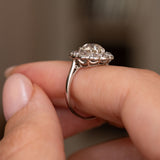 A close-up of a platinum Antique, Edwardian diamond cluster engagement ring shown worn on a person's finger from the side to display the ring's profile.