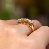 A close-up of a yellow gold Antique, Edwardian brown diamond three-stone engagement ring worn on a person's finger, set against a softly blurred green and brown background and shown from a low angle to display the claw settings and pierced gallery.