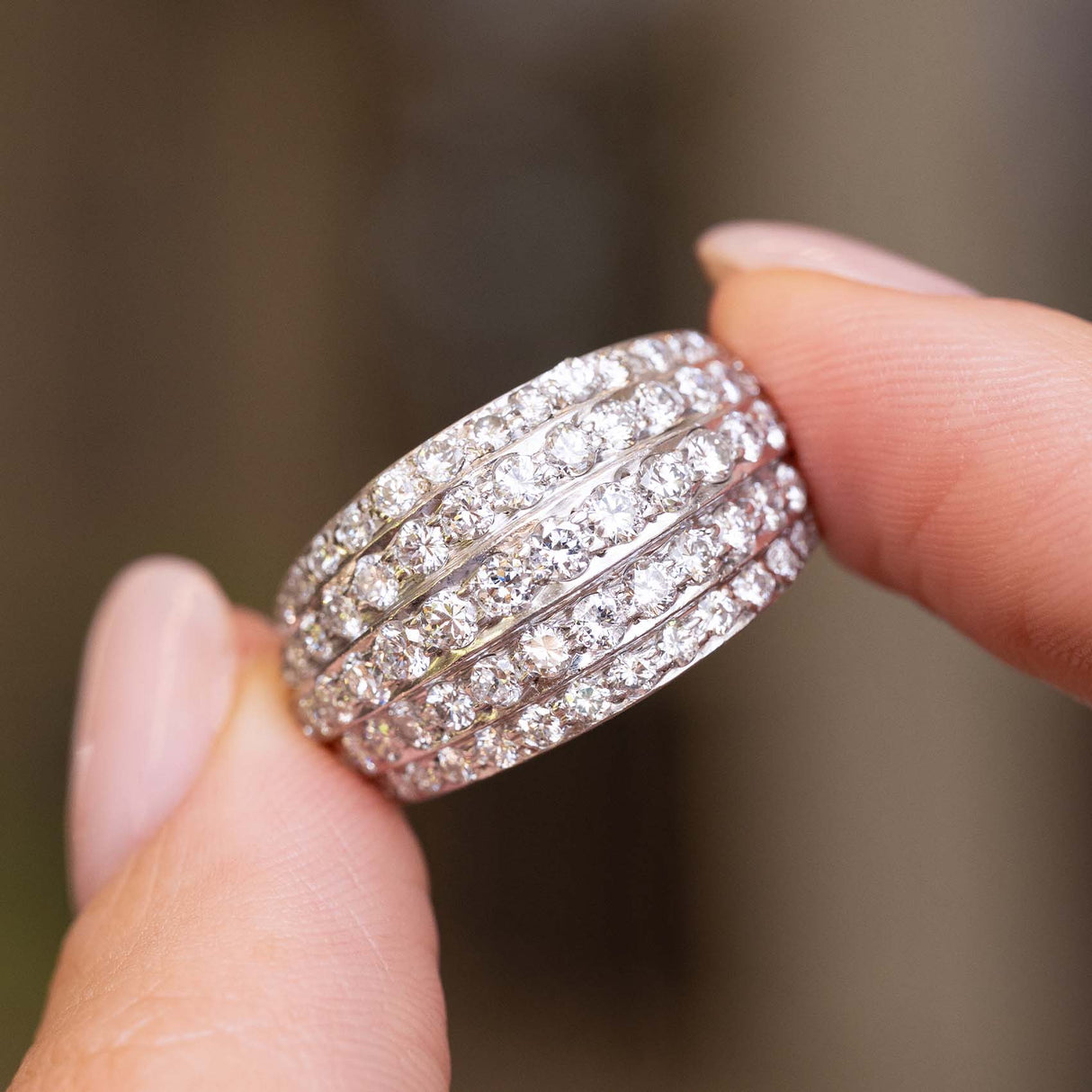 A close-up of A Vintage, 1950s diamond eternity bombe cocktail ring in white gold held between fingers, shown against a blurred neutral background.