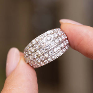 A close-up of A Vintage, 1950s diamond eternity bombe cocktail ring in white gold held between fingers, shown against a blurred neutral background.