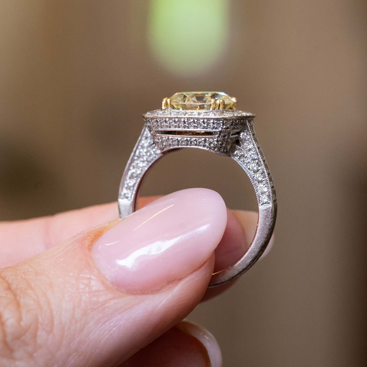 A close-up of a hand holding a white gold Vintage, 1990s yellow tinted diamond cluster engagement ring against a blurred background