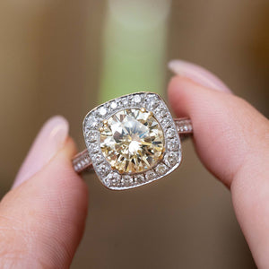 A close-up of a hand holding a Vintage 1990s cluster halo engagement ring with a large round yellow diamond stone and surrounding diamonds on a soft blurred background.