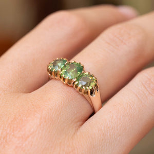 A close-up of a gold trilogy ring with three oval green zircons gemstones worn on a finger and seen from an angle of the side.