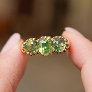 A close-up of a gold three-stone ring held between fingers featuring three green zircons against a blurred background.