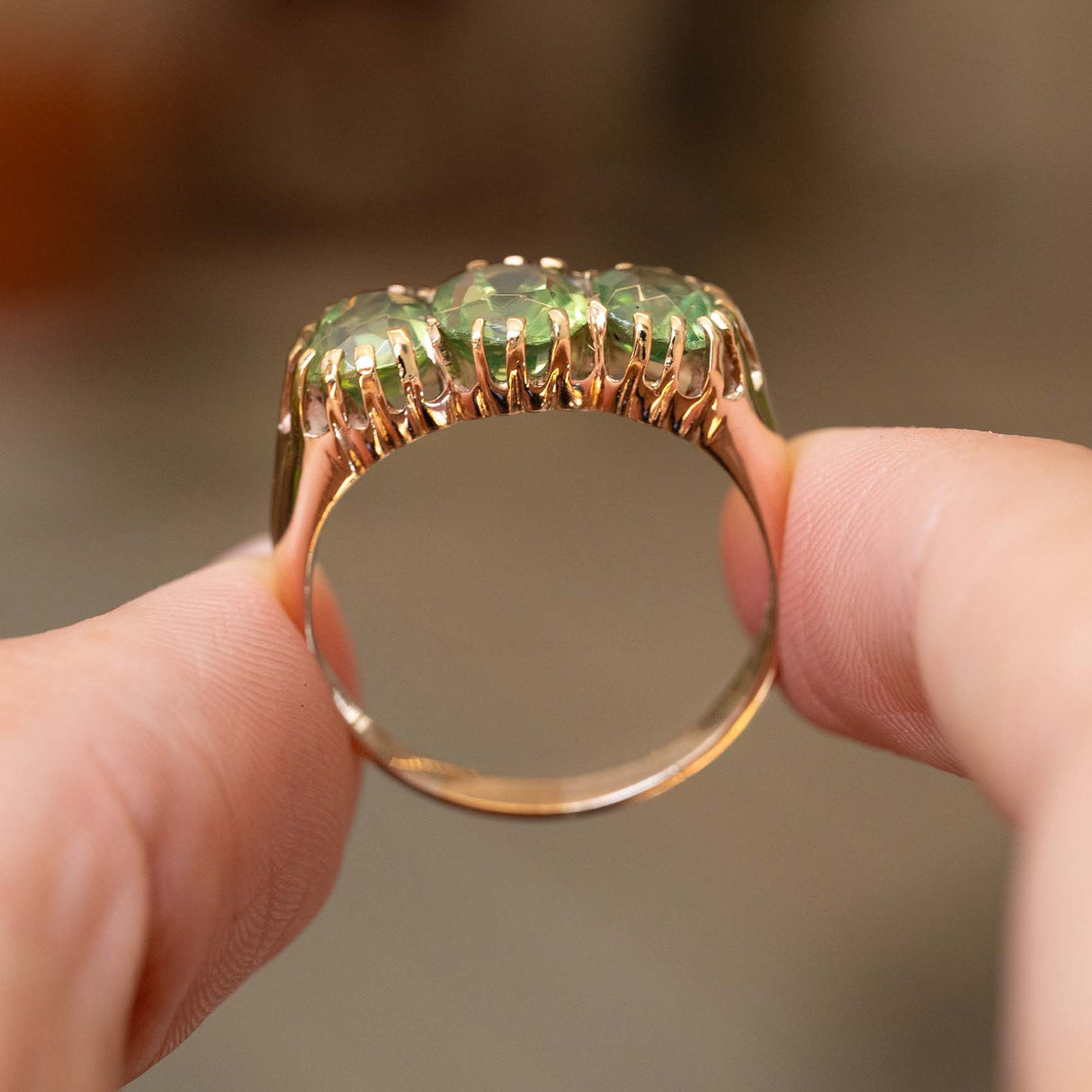 A close-up image of the profile of a gold three-stone ring held between fingers featuring three green zircons against a blurred background to show the band and setting of the ring.