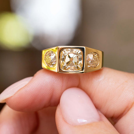 A close-up of a gold three-stone gypsy style diamond ring balanced on a finger against a softly blurred background. 