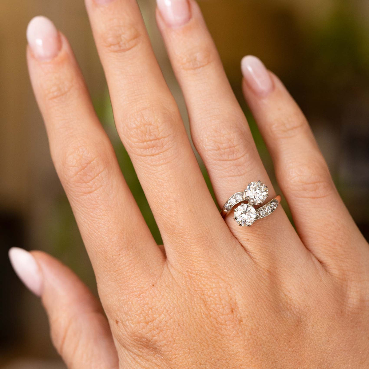 A close-up of a hand wearing a Antique, Edwardian diamond toi et moi engagement ring in platinum and gold set against a soft, blurred background.