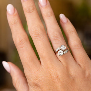A close-up of a hand wearing a Antique, Edwardian diamond toi et moi engagement ring in platinum and gold set against a soft, blurred background.