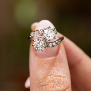 Fingers holding up a diamond two stone engagement ring in yellow gold antique Edwardian held up to the camera to show the details of the diamonds.
