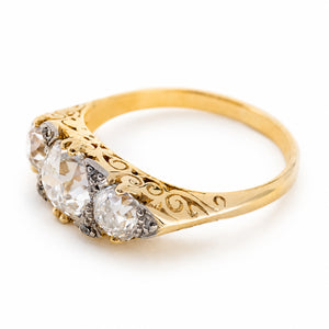 Angled side view of an antique three-stone engagement ring highlighting the open-back diamond settings and ornate scrolling detail carved into the 18ct yellow gold gallery and shoulders.