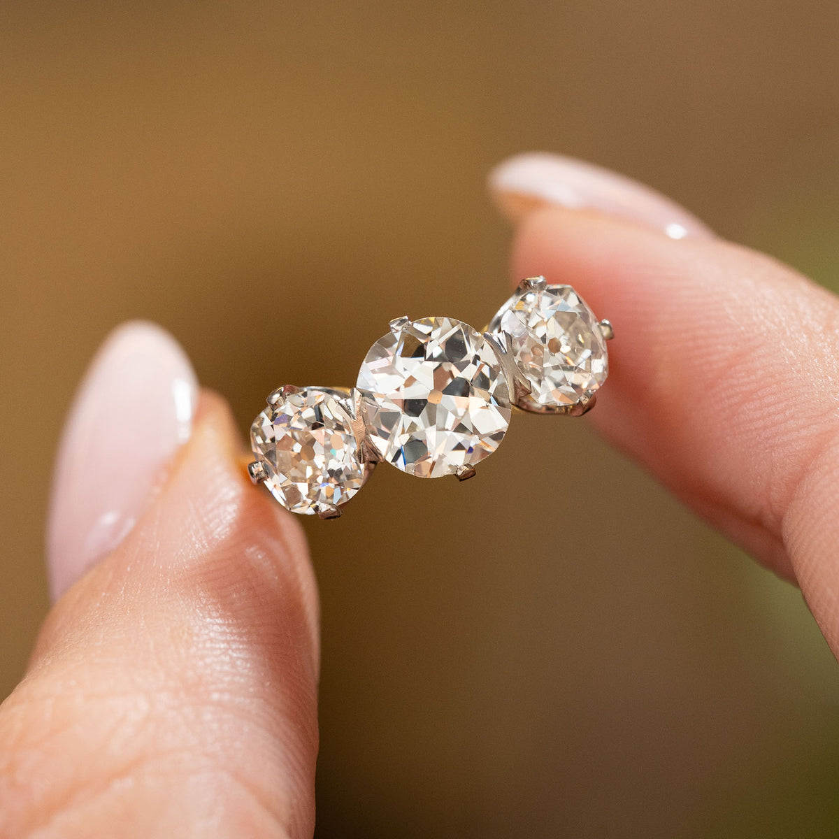 A close-up of an Antique, Edwardian old-cut diamond three-stone engagement ring held in between two fingers against a softly blurred background.