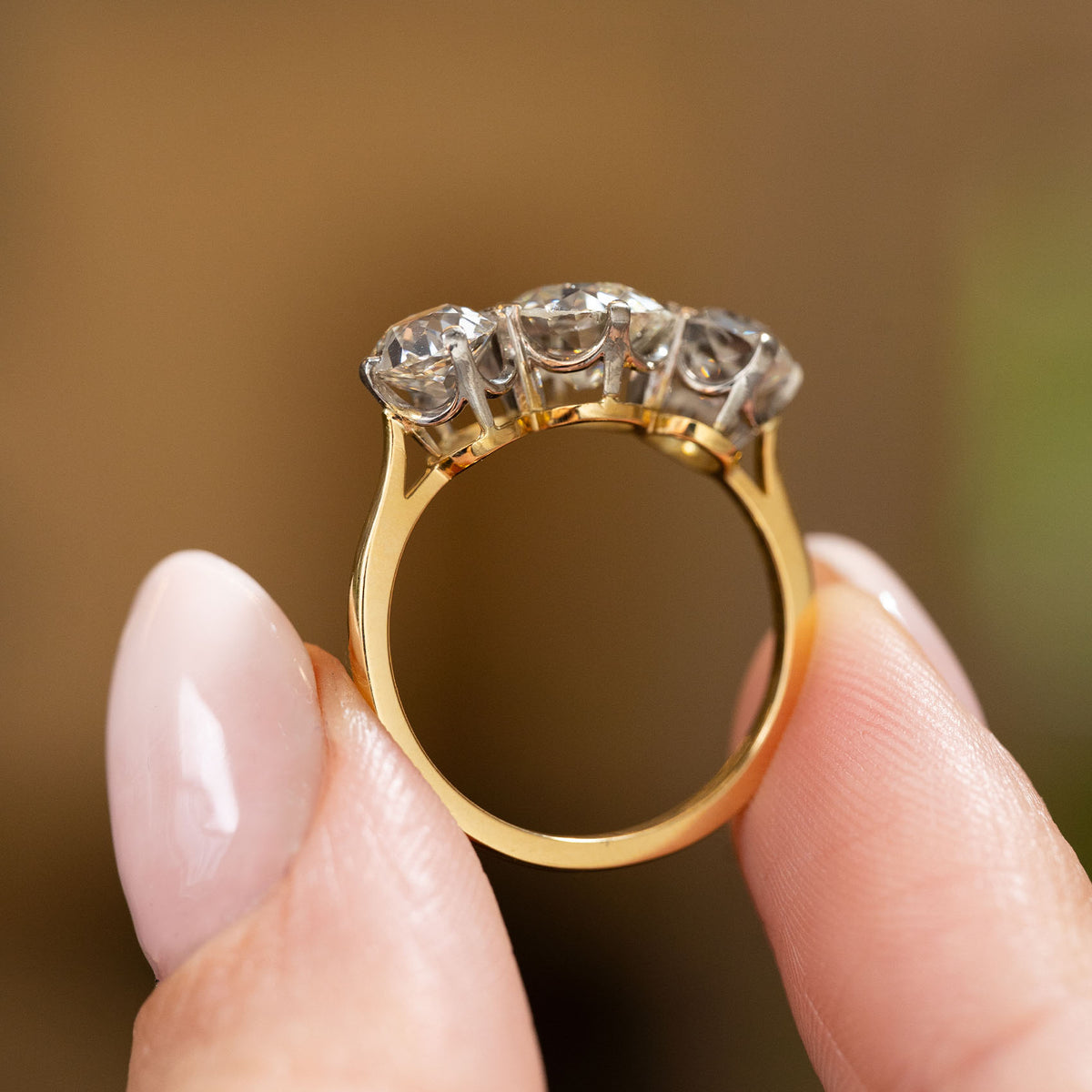 A close-up image of two fingers holding a antique Edwardian era diamond trilogy engagement ring with a yellow gold band. The ring is being held to show off its profile and shank.