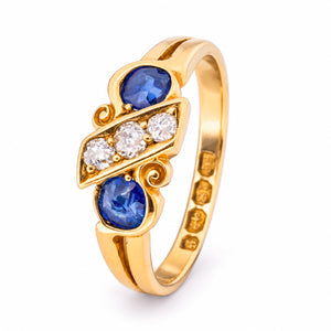 A raised three-quarter perspective reveals the diagonal diamond panel, opposing blue sapphires and sculptural scroll motifs.