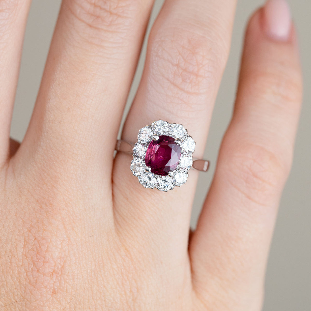 Close-up of the ruby cluster ring worn on a hand, highlighting the oval gemstone and scalloped arrangement of surrounding diamonds.