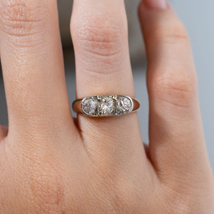 Close-up of an antique Edwardian diamond three-stone engagement ring worn on a finger, featuring three round old-cut diamonds in a platinum setting with a smooth yellow gold surround.