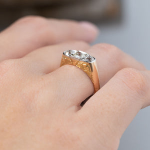 Side angle of an antique Edwardian diamond three-stone engagement ring worn on a finger, revealing its low-profile platinum diamond setting and decorative engraved yellow gold shoulders.