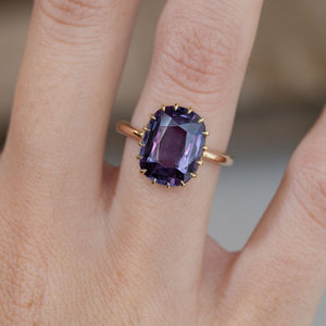 Close-up of the rectangular purple sapphire solitaire ring worn on a finger, featuring an intricate yellow gold claw setting.
