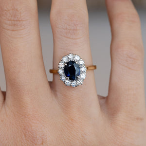 Close-up of the oval dark blue sapphire framed by brilliant-cut white diamonds, worn on a finger.